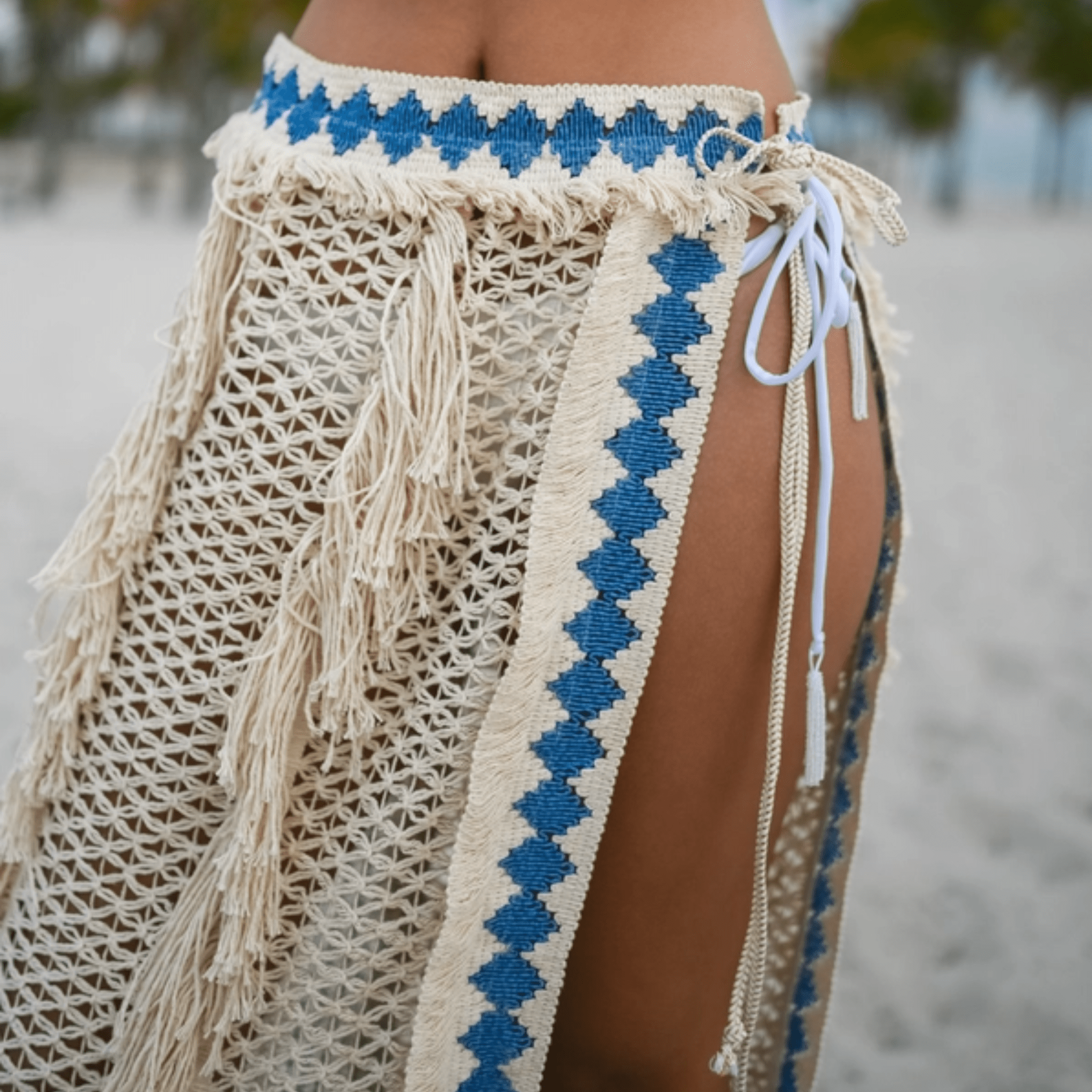 A lightweight resort knit pareo made of soft, breathable cotton, featuring a delicate knit pattern, natural fringes, and a blue embroidered border. Designed with a flattering tie-waist, this bohemian wrap moves gracefully—ideal for beach days or festival nights.
